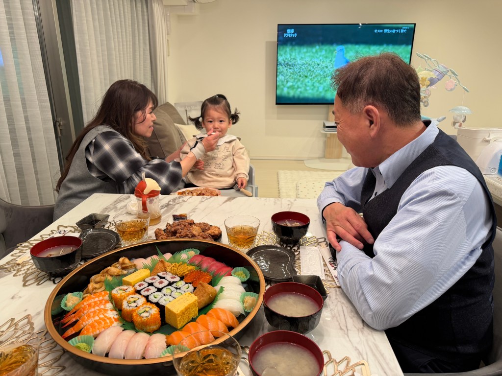 テーブルで寿司を楽しむ家族の写真。女性が子供に食べ物を与えており、男性が微笑んでいる。テレビでは何かが放送されている。
