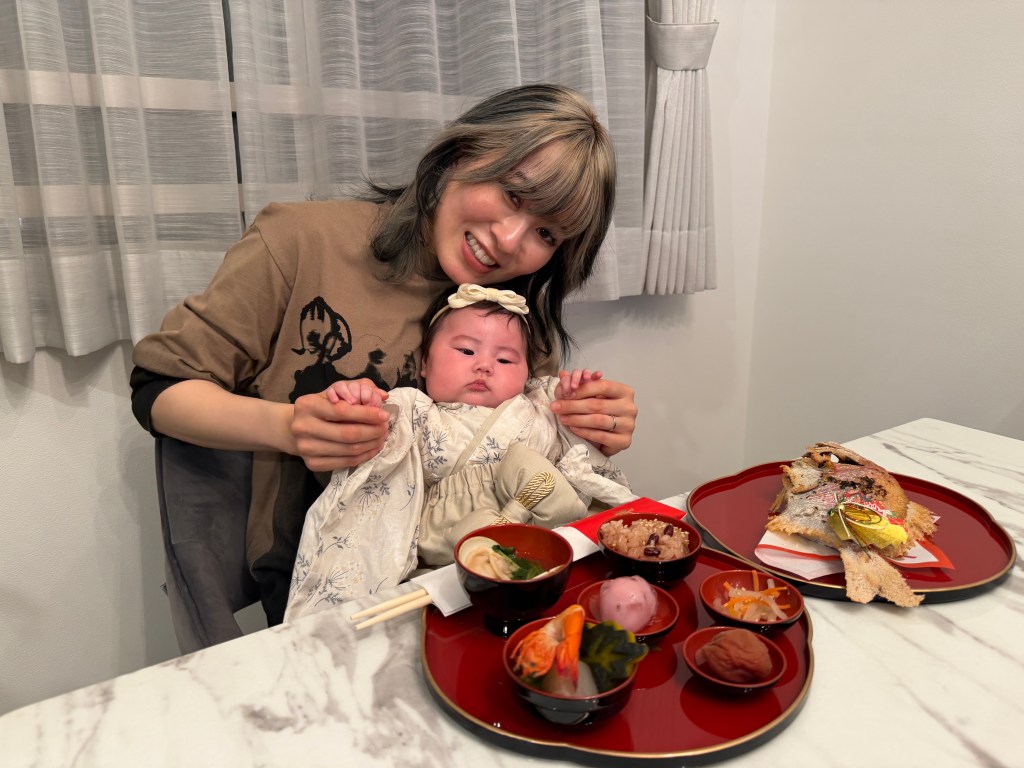 赤ちゃんと笑顔の女性が座っている。テーブルの上には豪華な和食のセットが置かれている。