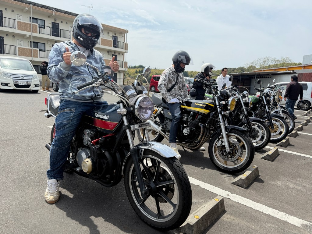 バイクに乗っている男性がサムズアップをしている。周囲には複数のバイクが並んでおり、背景には建物が見える。