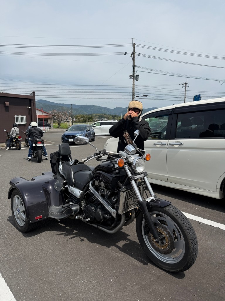 黒いバイクとサイドカーに乗る人物がカメラを構えている様子。背景には他のバイクや車、山々が見える。