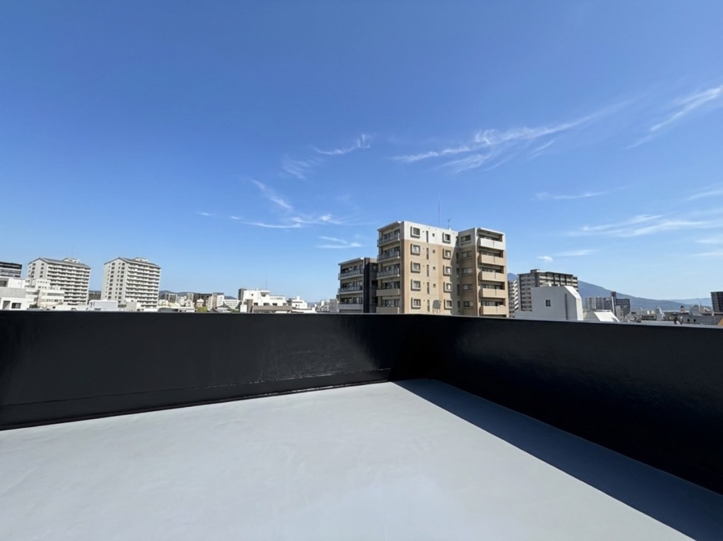 青空の下、ビル群の景色が広がる屋上の風景。手前には黒いバルコニーの手すりが見える。