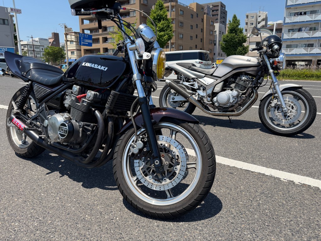 二台のバイクが並んでいる写真。手前には黒いカワサキのバイク、奥にはシルバーのバイクが見える。背景には建物が並ぶ街の風景が広がっている。